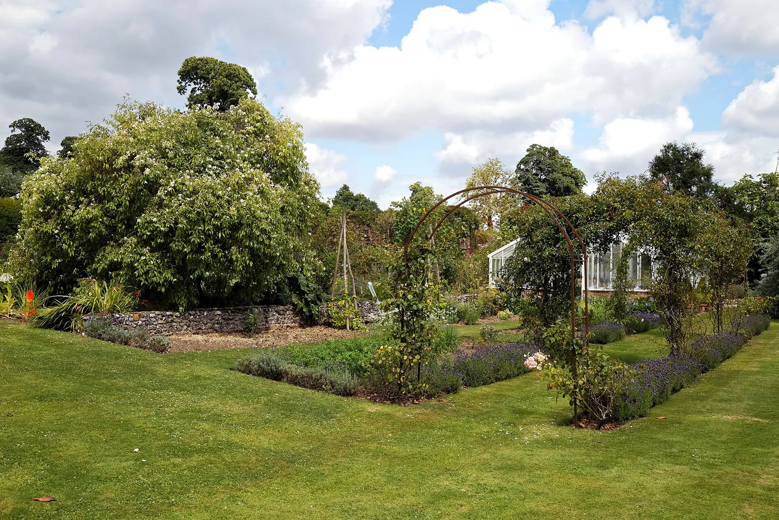 A garden lawn with a rose trellis and old trees under a summer sky