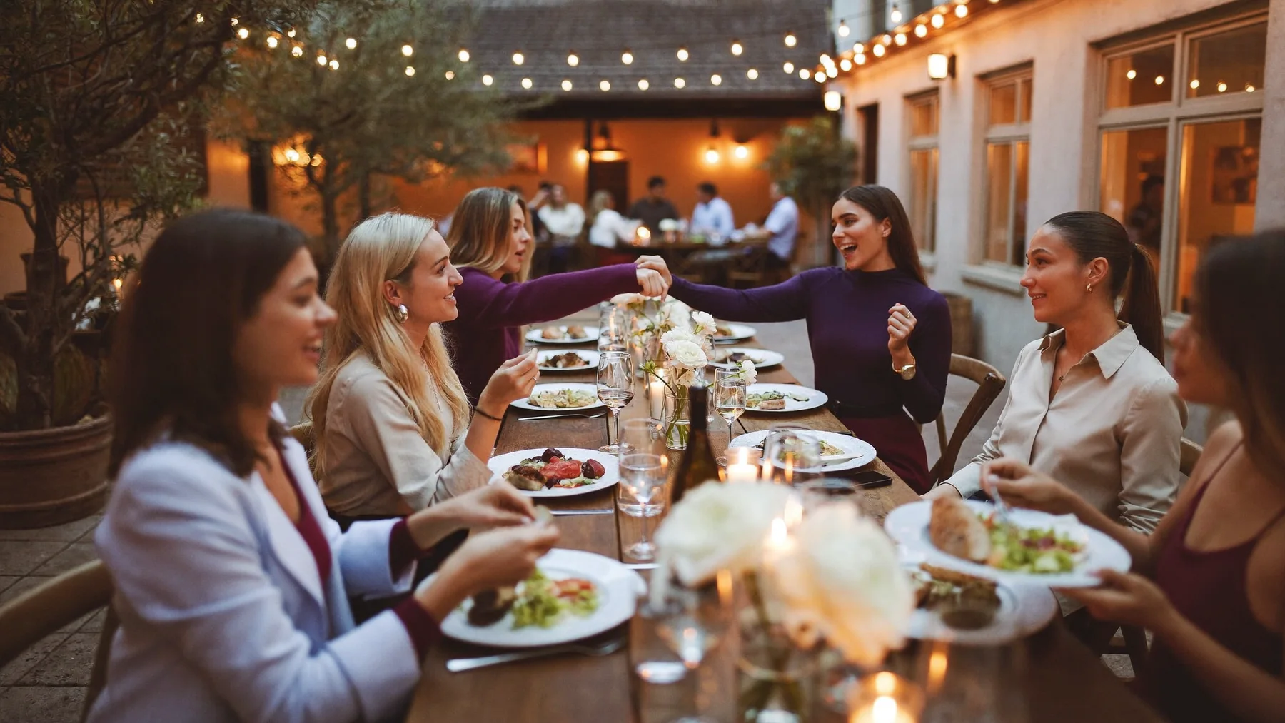 Women laughing around a long candlelit table in a courtyard under string lights