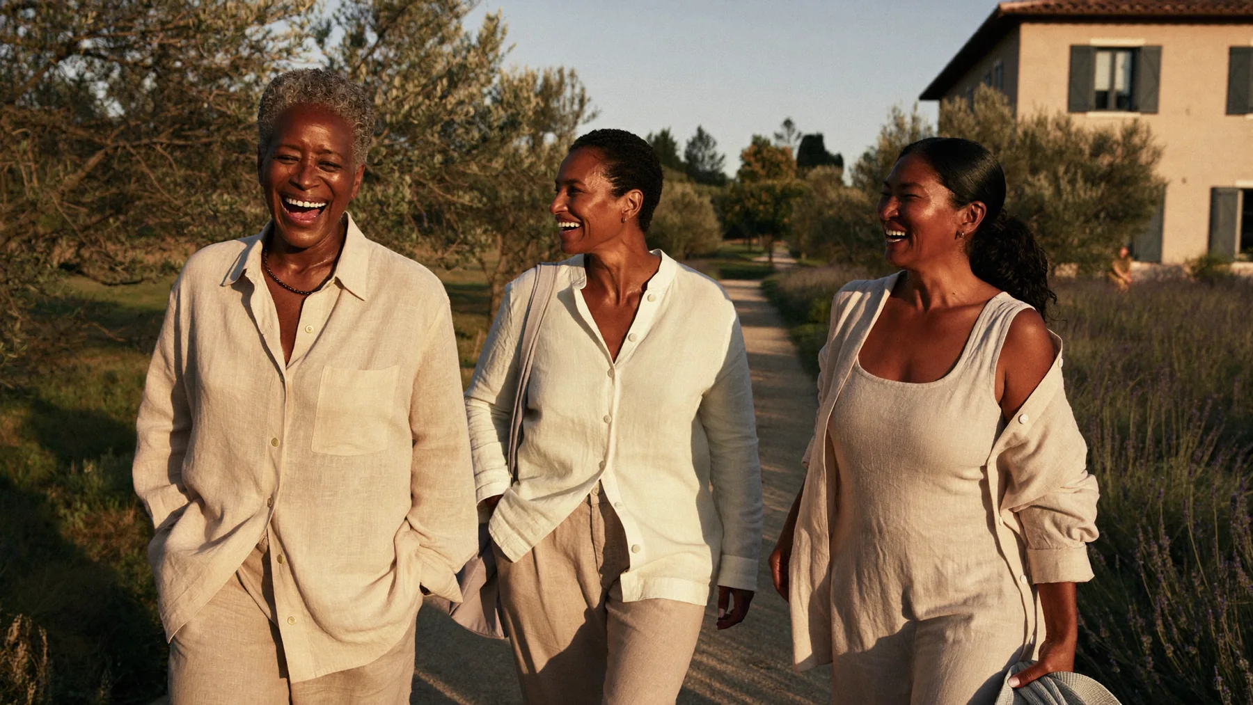 Three women in linen laughing as they walk along a garden path at a villa at golden hour